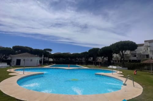 Piscina circular azul brillante en el resort Barrosa Home con tumbonas, césped verde y pinos mediterráneos bajo cielo despejado