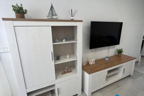Moderna sala de estar en Barrosa Home con muebles blancos, estantes de madera, TV montada en pared y decoración costera