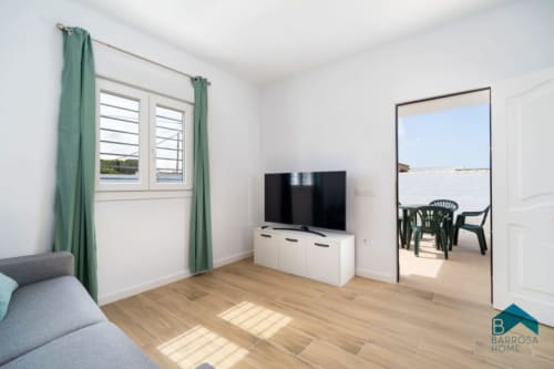 Modern living room at Barrosa Home with TV, green curtains, wooden floor, and ocean view access through open door