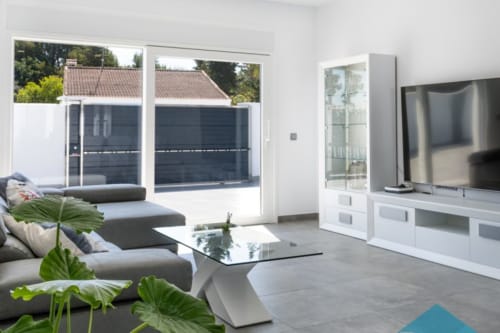 Sala de estar moderna en Barrosa Home con sofá gris, mesa de cristal, televisor y amplias ventanas con vistas a terraza y jardines.