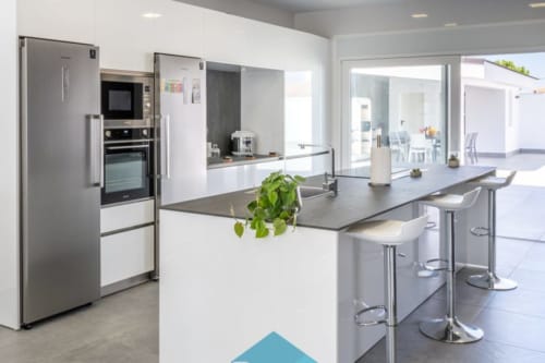Cocina moderna en Barrosa Home con electrodomésticos de acero inoxidable, muebles blancos, isla con encimera oscura y asientos en barra