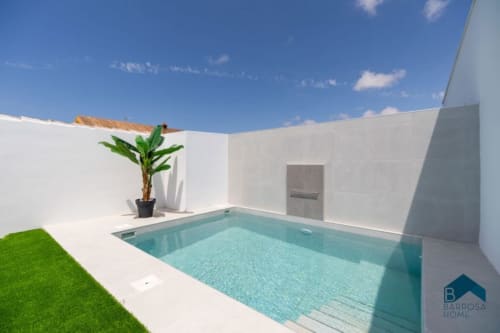 Patio moderno blanco con piscina turquesa, césped verde, maceta y cielo azul en Barrosa Home, España