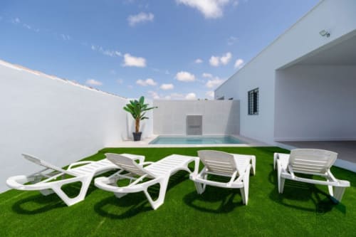Jardín trasero de Barrosa Home con tumbonas blancas, piscina, césped artificial y edificio moderno blanco bajo cielo azul