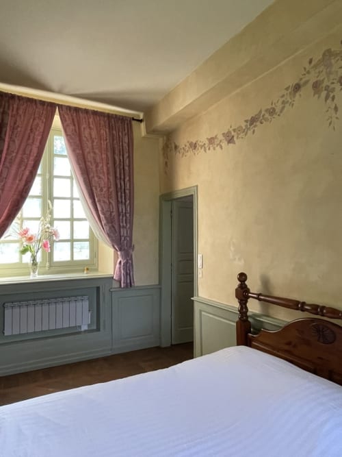 Chambre du Gîte Voltaire au Château de la Motte : lit en bois, murs crème, rideaux bordeaux et vue sur le jardin