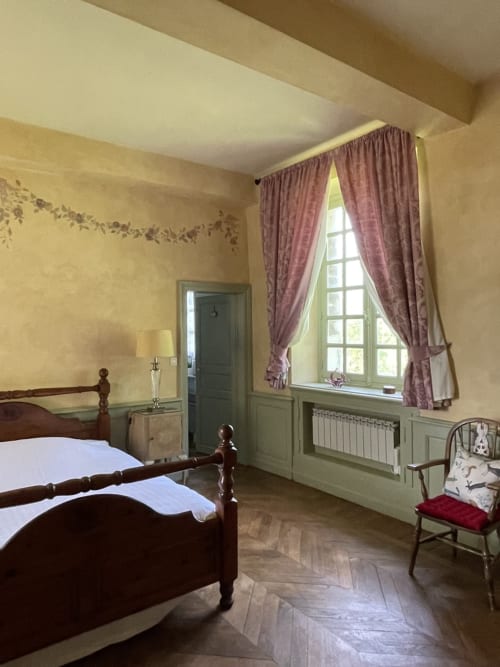 Chambre Gîte Voltaire au Château de la Motte avec lit en bois, murs jaunes, rideaux bordeaux et vue sur le jardin