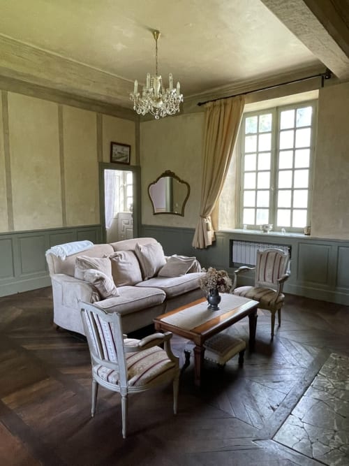 Élégant salon du Château de la Motte avec canapé crème, fauteuils d'époque, lustre en cristal et vue sur les jardins