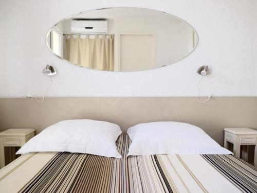 Modern hotel bedroom at Hôtel l'Orangeraie Lavandou with striped bedding, white pillows, oval mirror, and garden view.