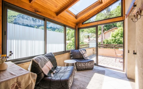 Bright sunroom with wooden beams, black seating, and panoramic mountain views of Millau, France from Dourbie Cottage