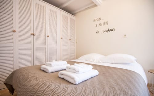 Dourbie Cottage bedroom with white pillows, folded towels on bed, beige quilt, white louvered wardrobe, French text wall art