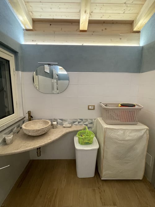 Bagno moderno di Villa Maria con specchio rotondo, piano in pietra, travi in legno, cesti per la biancheria e pareti piastrellate bianche
