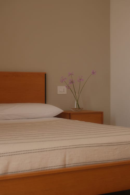 Bedroom at Nafura hotel in Raffadali with wooden bed, white pillow, purple flowers in glass vase on nightstand, and beige