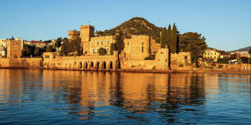 Medieval waterfront castle with golden stone towers reflecting in blue water, Spéracèdes France