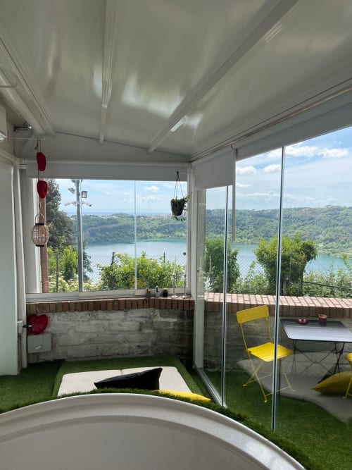 Sala de sol luminosa com vista para o lago na Casa Vacanze Abitare il tempo, Velletri. Grama sintética, cadeira amarela, plantas penduradas.