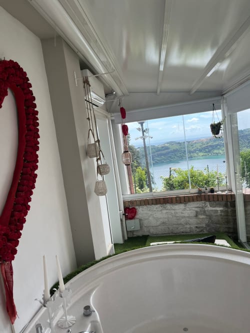 Banheiro luxuoso com banheira oval, guirlanda de flores vermelhas, cestos suspensos e vista panorâmica do lago em Velletri