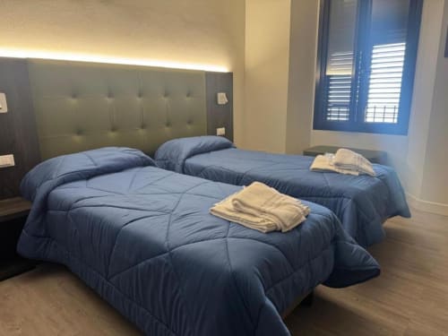 Camera quadrupla all'hotel NOI di Vergato con tre letti, coperte blu, testata imbottita, pavimento in legno e finestra con vista sulla città