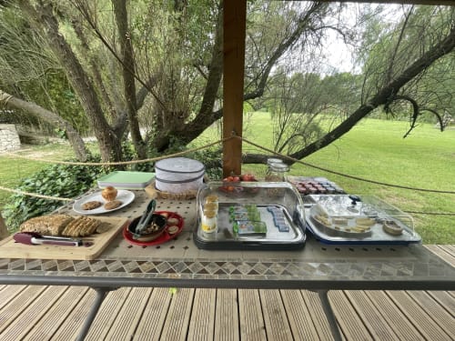 Pique-nique en plein air aux Jardins Du Soleil avec pain frais, fromage et vin, vue sur prairie verdoyante et saules.