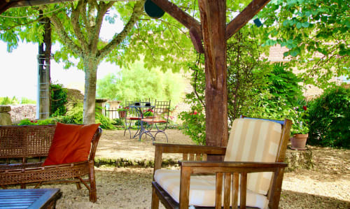 Patio ombragé du Gîte La Vieille Ferme à Saint-Geniès avec chaises en bois et coussins rouges, entouré de verdure luxuriante
