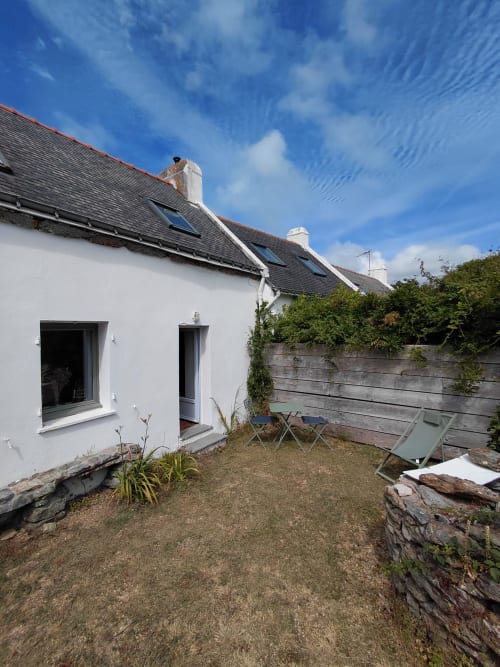 Chaumière blanche aux ardoises sombres, patio aménagé avec sièges, murs en pierre, ciel bleu dégagé - cottage Kergue'ch