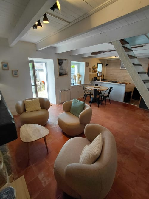 Salon cottage moderne avec fauteuils beiges cintrés, tables en bois et carrelage terracotta. Cuisine et salle à manger visibles