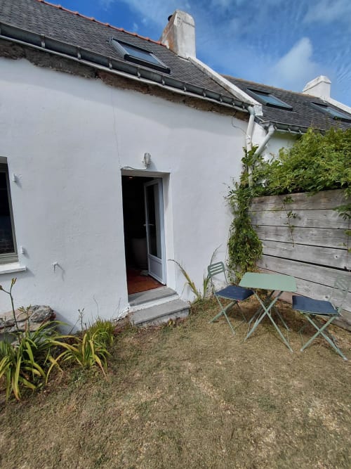 Cottage blanc avec toit ardoise et panneaux solaires, cour gravillonnée avec chaises de camping et bac en bois, vue sur le jardin