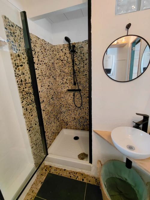 Salle de bain moderne avec douche en galets, miroir rond, lavabo blanc et robinetterie noire au gîte Kergue'ch