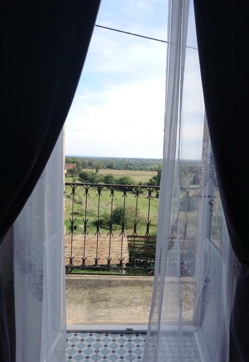 Des portes-fenêtres ouvertes avec voilages encadrent la vue champêtre sur les champs et forêts de la chambre Le chant de la cloche à La