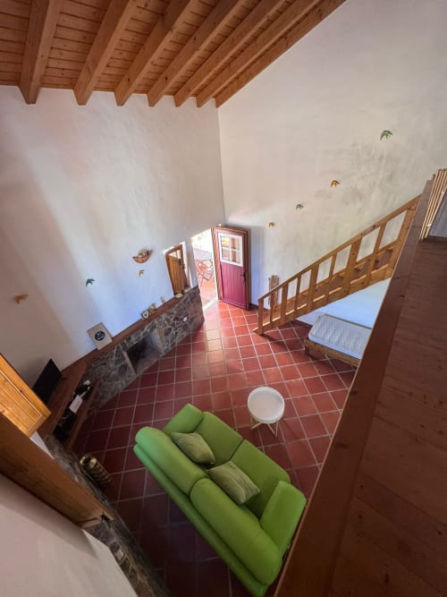 Quarto iluminado na T Monte B&B Porto Côvo com sofá verde, vigas de madeira, piso de azulejos vermelhos e acesso ao jardim