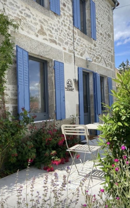 Cour fleurie de La Maison De Fleurat avec volets bleus, chaise blanche, plantes en fleurs et murs de pierre à Domeyrot
