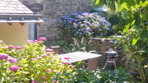 Charmante terrasse de jardin au Clos du Tay à La Gacilly avec hortensias fleuris, fleurs roses, table en bois et murs en pierre