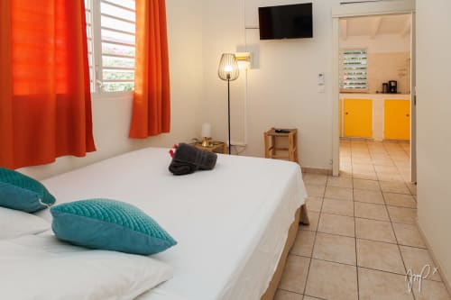 Chambre lumineuse du cottage caribéen Otantik avec coussins turquoise, lit blanc, rideaux oranges et vue sur les montagnes