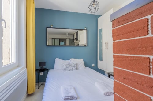 Chambre studio moderne à Metz avec mur d'accent bleu, mur de briques, lit, miroir et rideaux jaunes