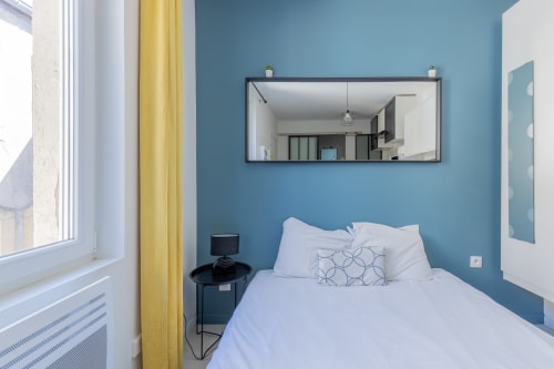 Chambre moderne à Metz aux murs bleus, lit blanc, rideaux jaunes et miroir au-dessus de la tête de lit