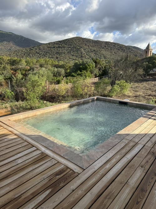 Piscine luxueuse avec terrasse en bois surplombant les collines boisées et le clocher lointain de Figari, Corse