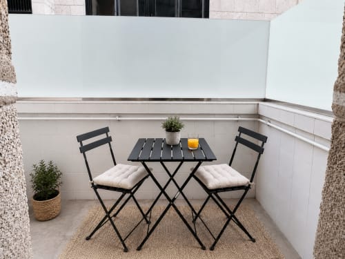 Zona de estar en el balcón del apartamento Antia Mare en Baiona con mesa, sillas negras y plantas en macetas con vistas a la ciudad.