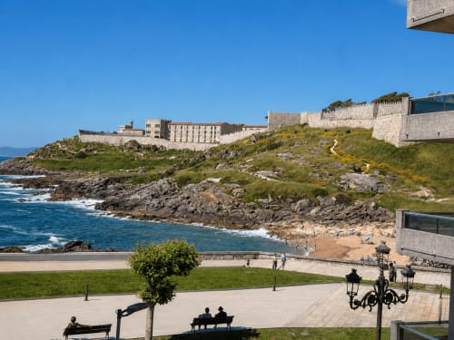 Vista costera de la fortaleza de Baiona con mar, playa y muros de piedra dominando ensenadas rocosas