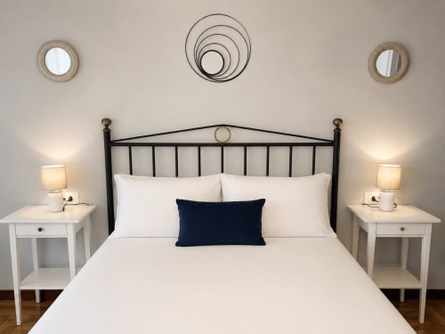 Moderno dormitorio del apartamento Holibai en Baiona con cama de hierro forjado negro, ropa de cama blanca, almohada azul marino y mesillas blancas
