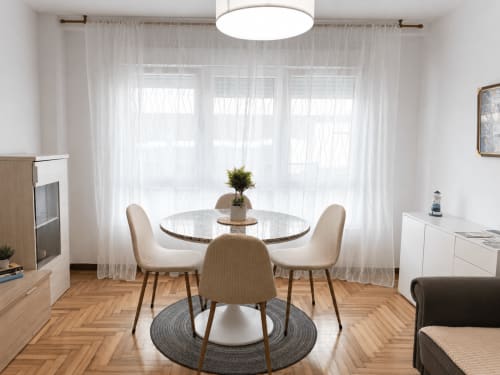 Luminoso comedor del apartamento Morriña en Holibai Baiona con mesa redonda, sillas beige y vistas al mar a través de cortinas translúcidas