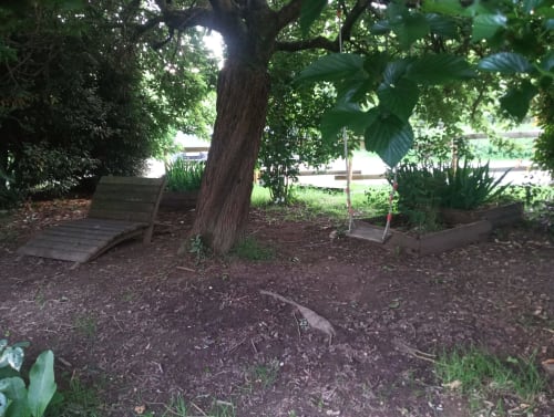 Coin ombragé du jardin de La Faerie cottage avec grand arbre, banc en bois, bacs de plantation surélevés et sol recouvert de paillis