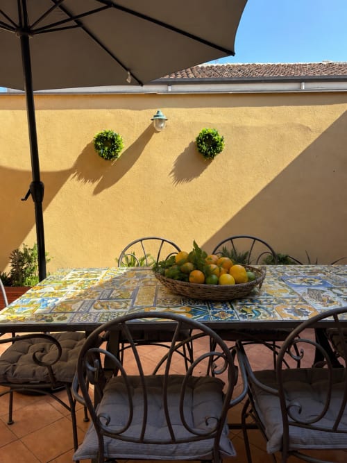 Soleggiato cortile siciliano con tavolo decorato in maiolica, sedie in ferro battuto, muro giallo, ombrellone e cesto di agrumi freschi