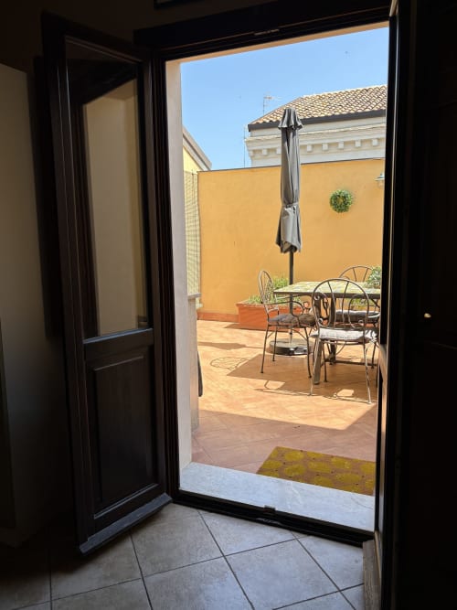 Porte nere aperte rivelano una terrazza soleggiata a Catania con tavolo da pranzo, sedie, ombrellone grigio e muro giallo circondato da