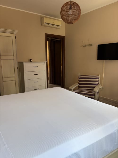 Ampia camera da letto presso Sicily In Home-Almarina a Catania con letto bianco, lampada a sospensione in vimini, TV, cassettiera e accenti a righe