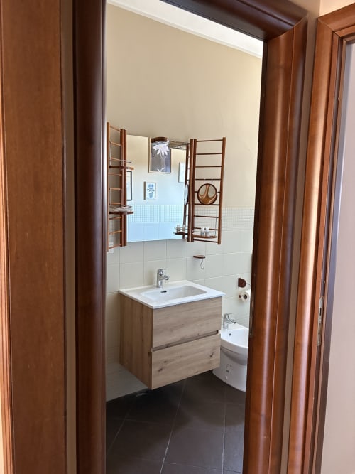 Bagno moderno con vanità in legno, specchio con ripiani, lavabo bianco, bidet e pareti piastrellate nell'appartamento In Home di Sicilia