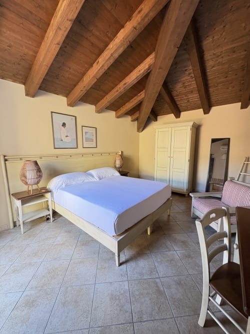 Camera luminosa con soffitto in travi di legno, letto matrimoniale, comodino bianco, armadio e pavimento piastrellato