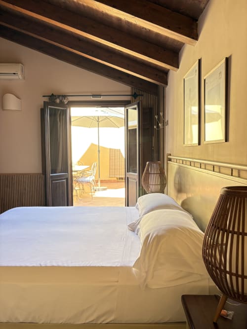 Camera da letto luminosa con letto bianco, travi in legno e porte aperte verso la terrazza soleggiata con ombrellone a Sicily In Home