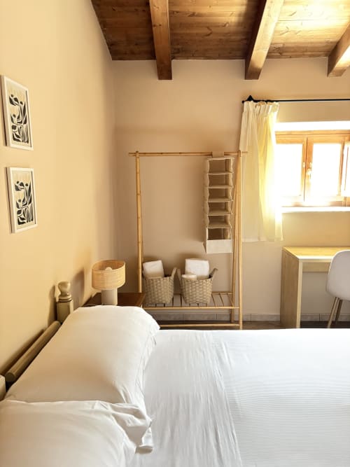 Luminosa camera da letto a Catania con travi in legno, biancheria bianca, scaffali e luce naturale dalla finestra