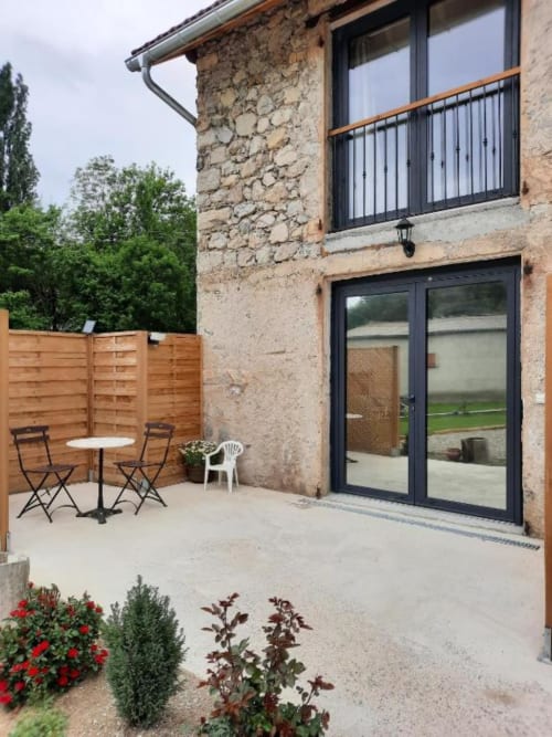 Patio du Petit Bégou avec bâtiment en pierre, portes-fenêtres noires, clôture bois et chaises de salle à manger à Saint-Auban-d'Oze