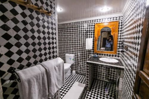 Salle de bain aux carrelages losanges noir et blanc, avec toilettes, lavabo, miroir doré et serviettes au Riad Fes Aicha