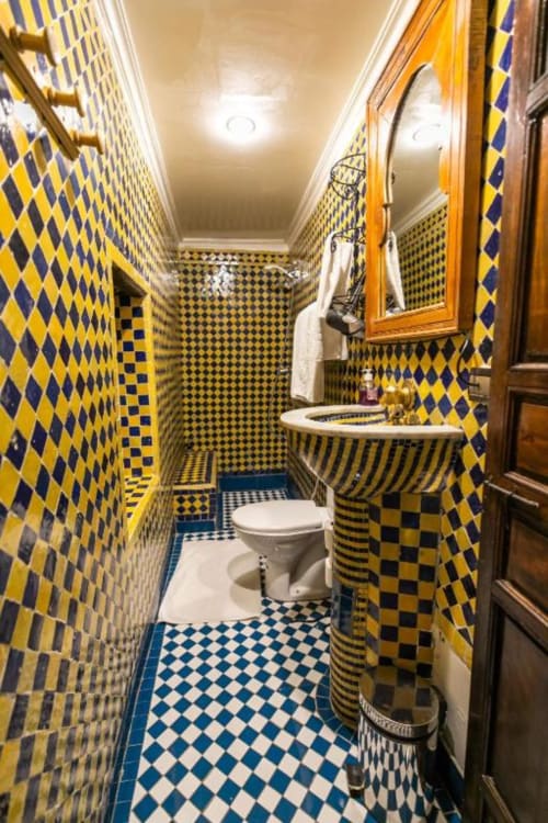 Salle de bain aux motifs géométriques jaunes et bleus du Riad Fes Aicha à Fès avec lavabo, toilettes et décor marocain traditionnel