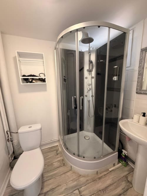 Salle de bain moderne dans l'appartement Kawichimmo à Louvres, avec douche vitrée incurvée, toilettes blanches et sol imitation bois.