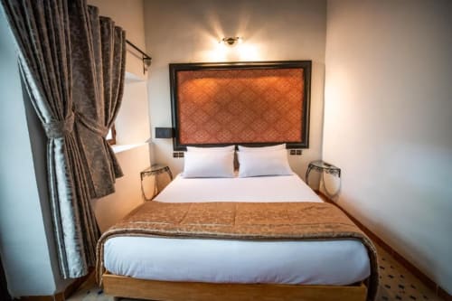 Chambre double douillette du Riad Dar Tahri à Fès : draps blancs, tête de lit orange, rideaux marron, lumière chaleureuse
